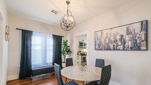 A dining area with a round marble table, black upholstered chairs, a cityscape painting, and a view into the kitchen. - Charming private house sleeps 4 with a large fenced yard, fast WiFi, smart TVs, and ample parking—near the Heights & Downtown. Features a primary king bed for a comfortable stay. (Houston)
