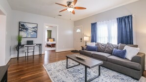 A modern living room with a grey sofa, a coffee table, a ceiling fan, and a view of a bedroom through an open door. - Charming private house sleeps 4 with a large fenced yard, fast WiFi, smart TVs, and ample parking—near the Heights & Downtown. Features a primary king bed for a comfortable stay. (Houston)