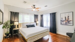 A bedroom with a bed, a ceiling fan, a nightstand, a lamp, a potted plant, and a painting on the wall. - Charming private house sleeps 4 with a large fenced yard, fast WiFi, smart TVs, and ample parking—near the Heights & Downtown. Features a primary king bed for a comfortable stay. (Houston)