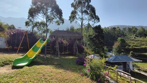 A playground with a green and yellow slide, a small wooden structure, and a fenced area with flowers. - Moon Wings Mountain Resort (Devikolam)