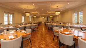 A spacious banquet hall with multiple tables set for dining, elegant chandeliers, and large windows allowing natural light.