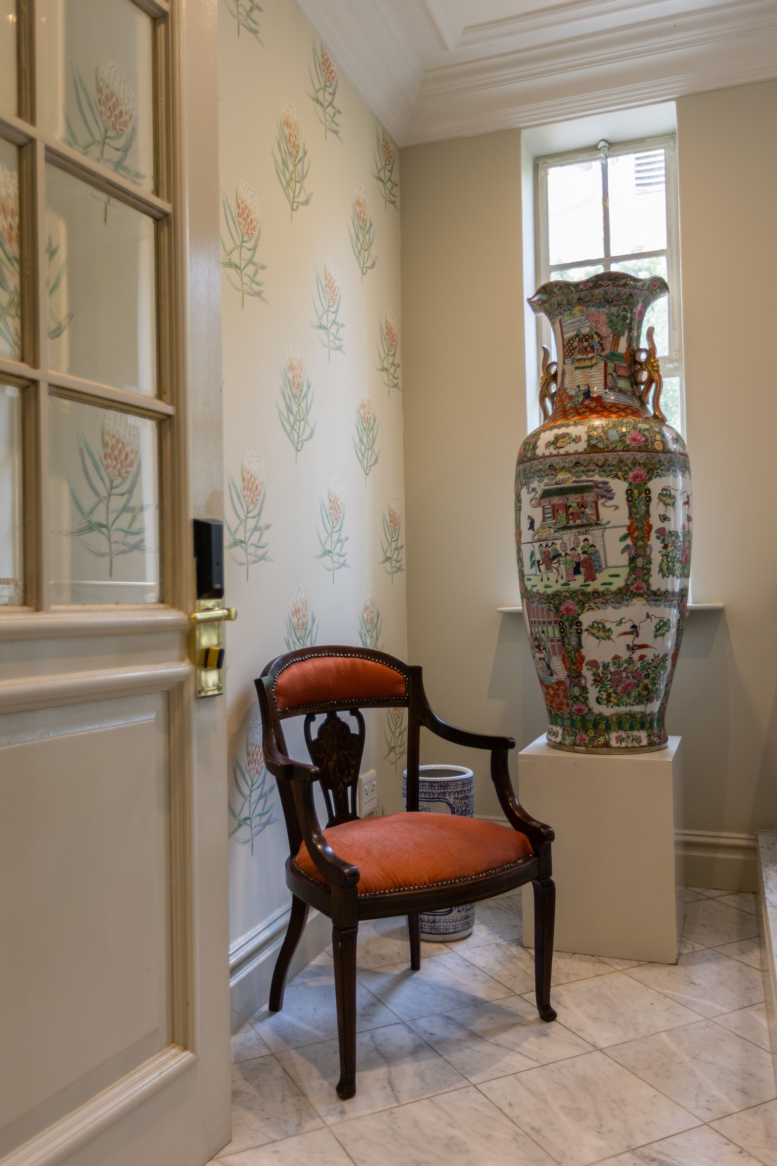 A room with a large decorative vase on a pedestal, a wooden chair with a cushion, and a door with frosted glass panels.