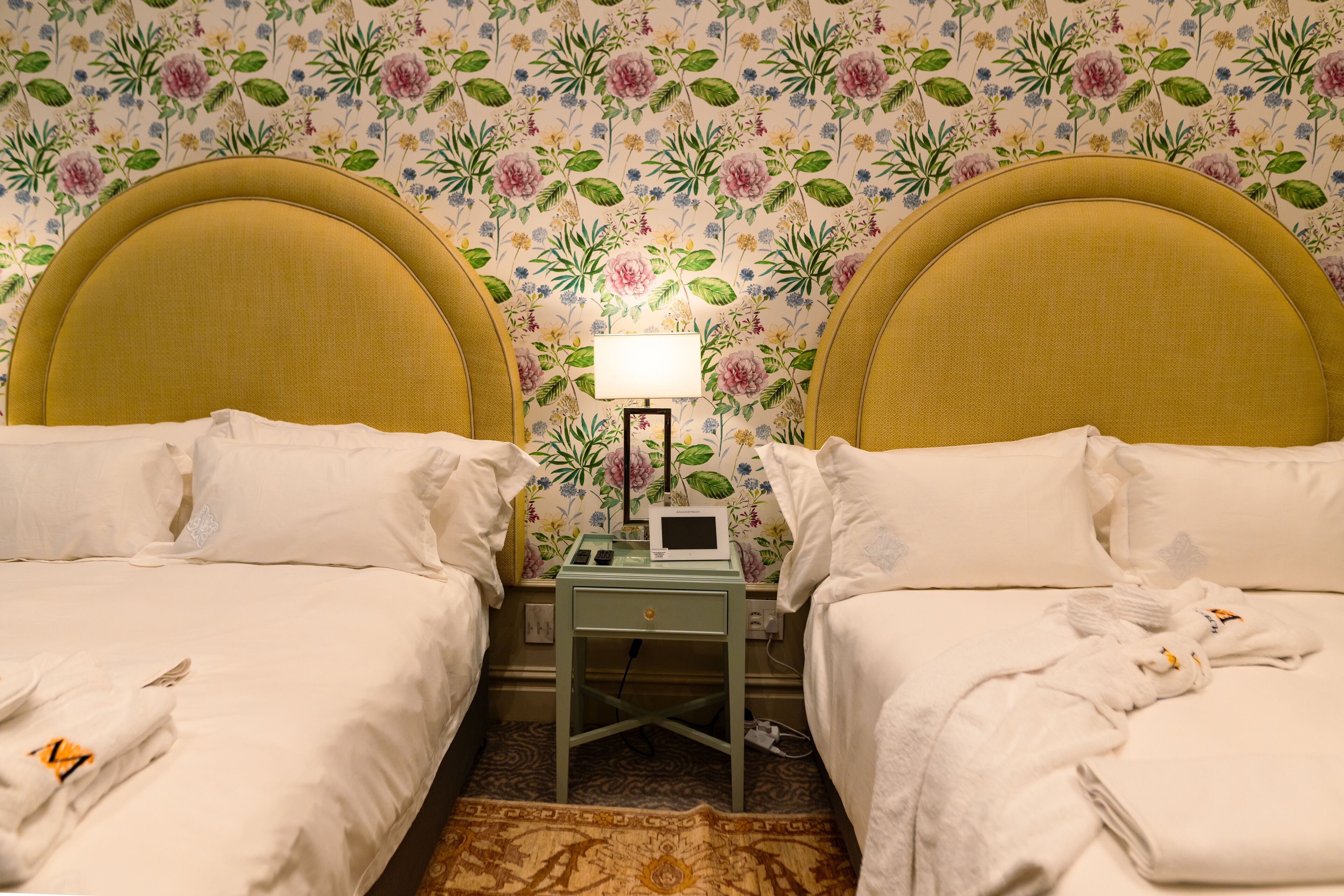 A hotel room with two beds, a nightstand with a lamp, and a floral wallpaper.