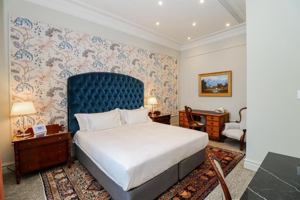 A hotel room with a large bed, two bedside tables with lamps, a desk with a chair, a patterned carpet, and a floral wallpaper.