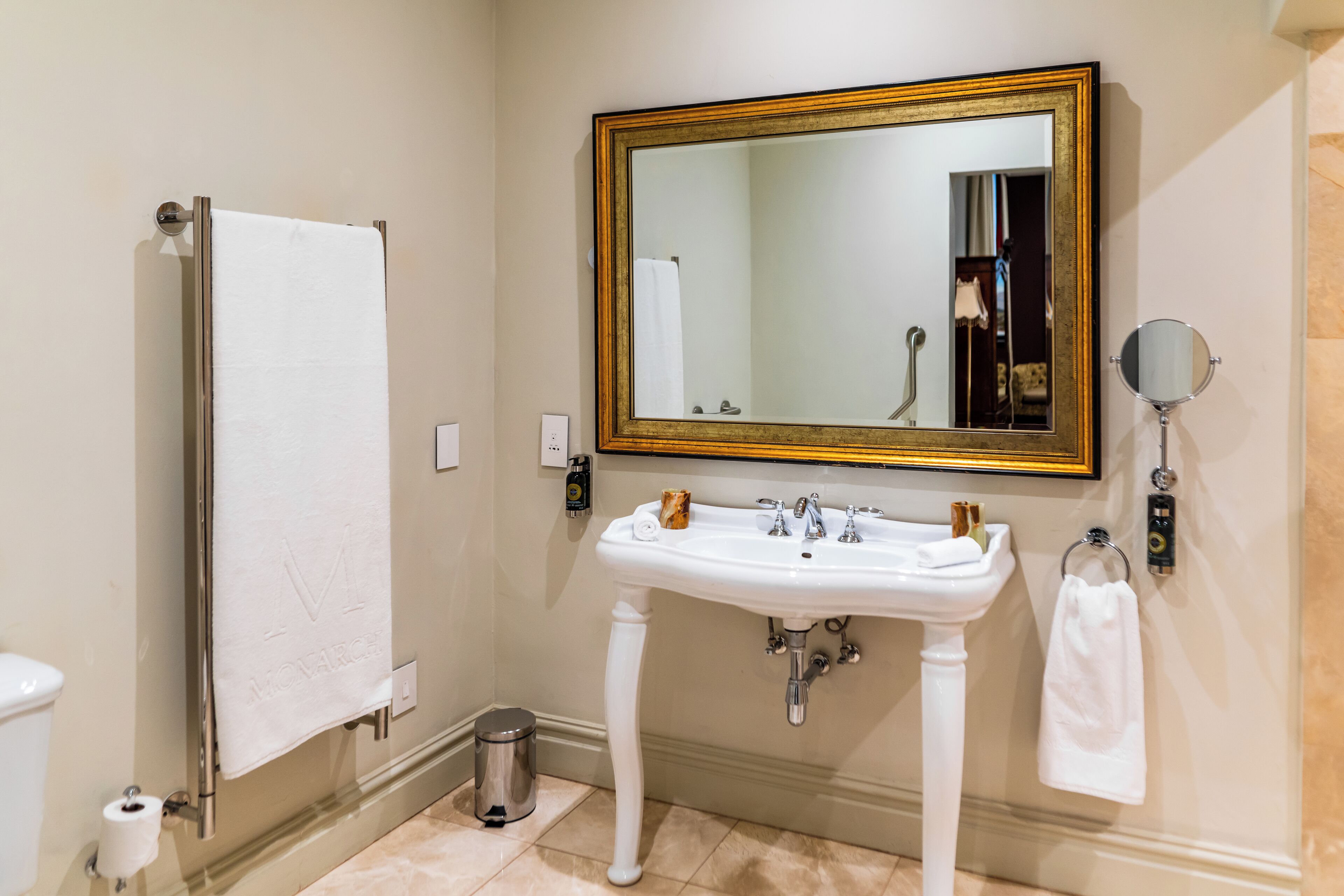 Un baño con un lavamanos pedestal, un espejo grande con marco dorado, un perchero y un bote de basura.
