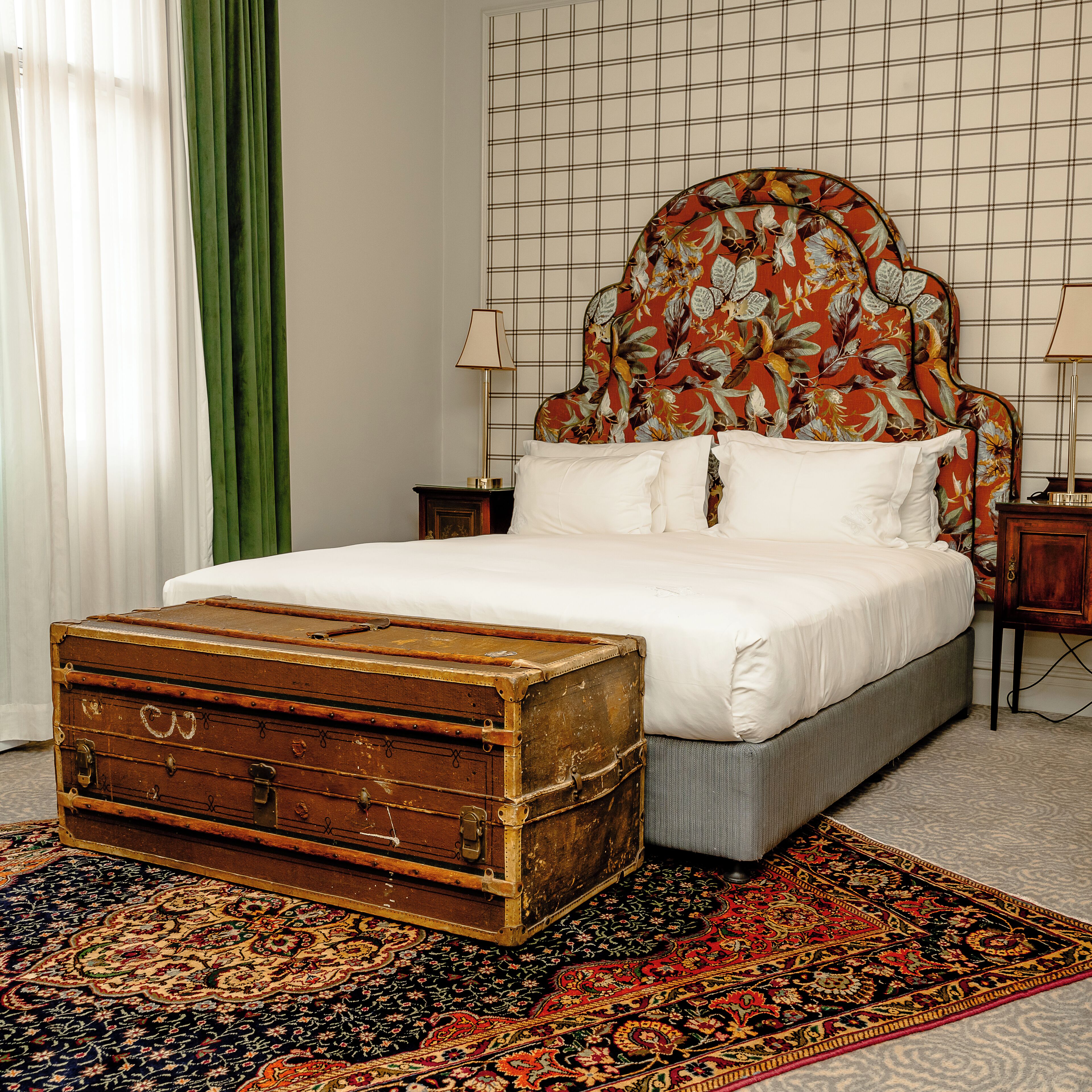 Un dormitorio con cama, mesita de noche, lámpara, cortina verde y maleta grande.
