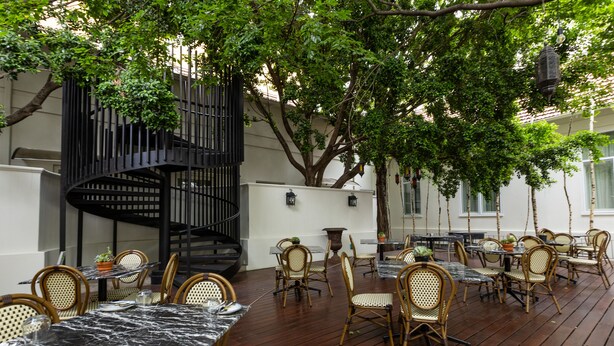 An outdoor dining area with wooden tables and chairs, surrounded by trees and a spiral staircase.
