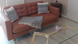 A living room with a brown sofa, two pillows, a knitted throw, a glass coffee table with a jar, and a framed picture on the wall.