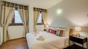 A hotel room with a large bed, two windows with curtains, a bedside table with a lamp, and a radiator.