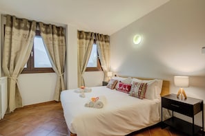Une chambre d’hôtel avec un grand lit, deux fenêtres avec des rideaux, une table de chevet avec une lampe et un radiateur.