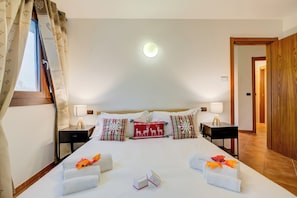 Een netjes opgemaakt bed met decoratieve kussens, een nachtkastje met lamp en uitzicht op de gang.