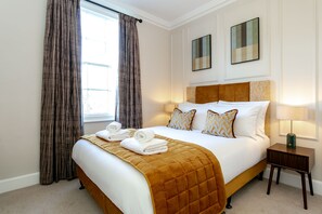 Una cama bien hecha con un edredón marrón, dos lámparas de noche y una ventana con cortinas.