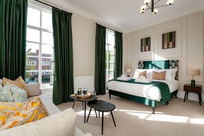 A hotel room with a large bed, a sofa, a small table, and a view of buildings outside.