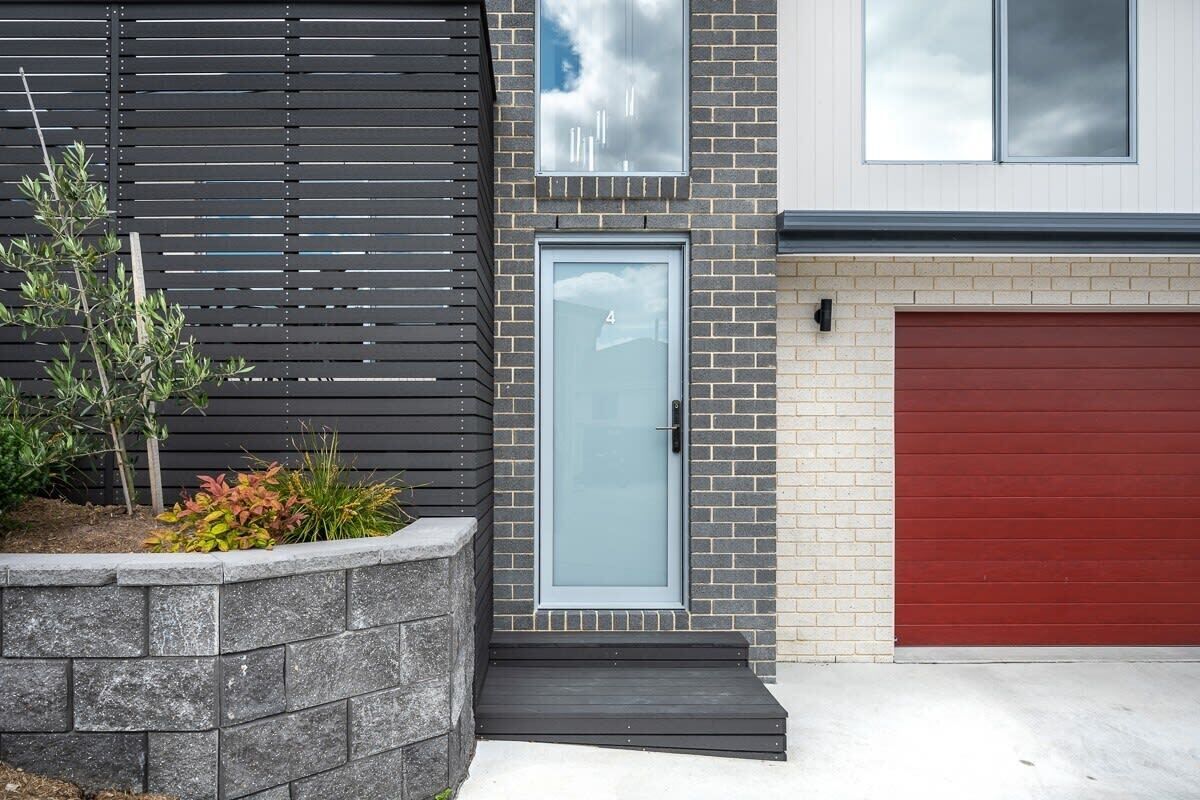 Een modern huis met twee verdiepingen, een gevel van grijze bakstenen, een rode garage deur en een kleine voortuin met planten.