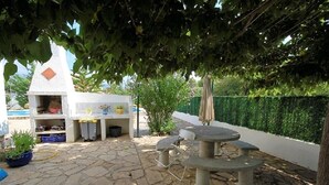An outdoor patio with stone flooring, a stone table with chairs, a small white structure with shelves, and a green hedge. - SAONA VILLAS (VN LARA): villa 8 people, private pool, wifi (Les Tres Cales)