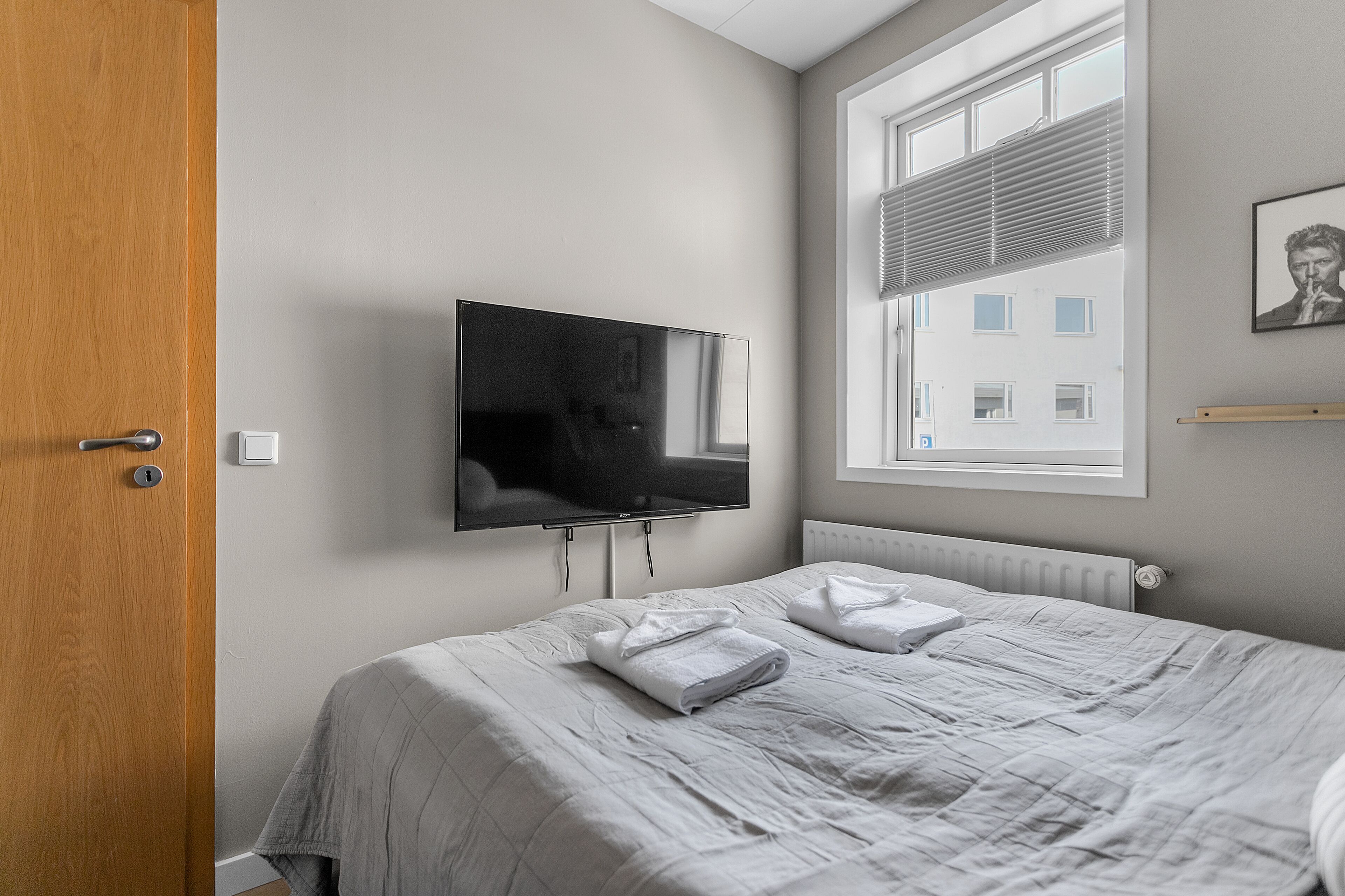 Ein Hotelzimmer mit Bett, einem flachbildfernseher an der Wand, einem Fenster mit Jalousien und einer Tür.