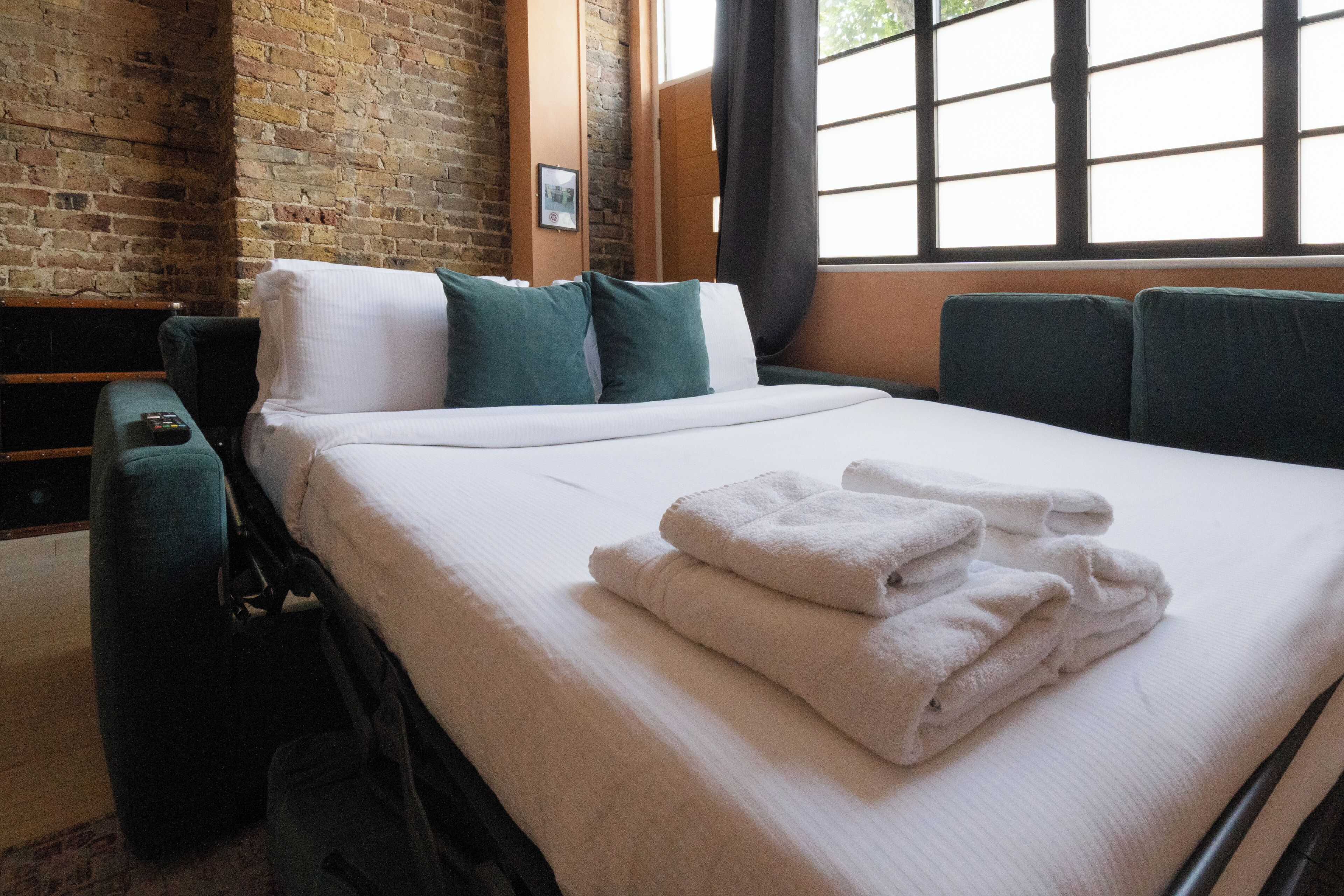 A neatly made bed with white linens and folded towels, a green upholstered headboard, and a brick wall.