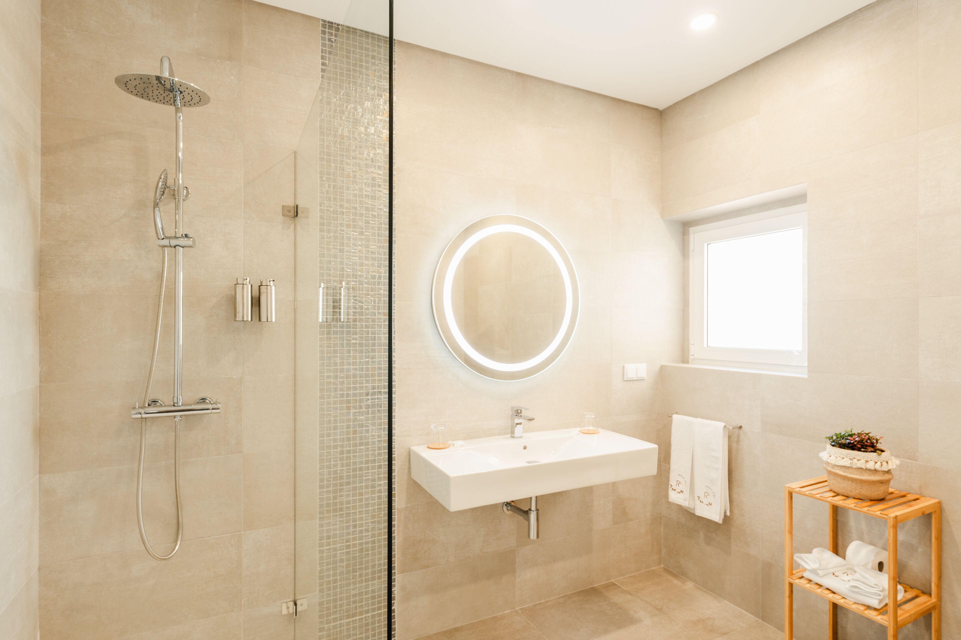 Une salle de bain moderne avec une douche à parois de verre, un miroir rond avec un éclairage intégré, un lavabo blanc et une petite étagère en bois sur laquelle est posée une plante.