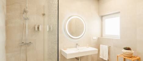 Une salle de bain moderne avec une douche à parois de verre, un miroir rond avec un éclairage intégré, un lavabo blanc et une petite étagère en bois sur laquelle est posée une plante.