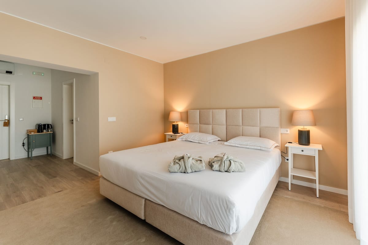 A hotel room with a large bed, two bedside tables with lamps, a small nightstand, and a door leading to another room.