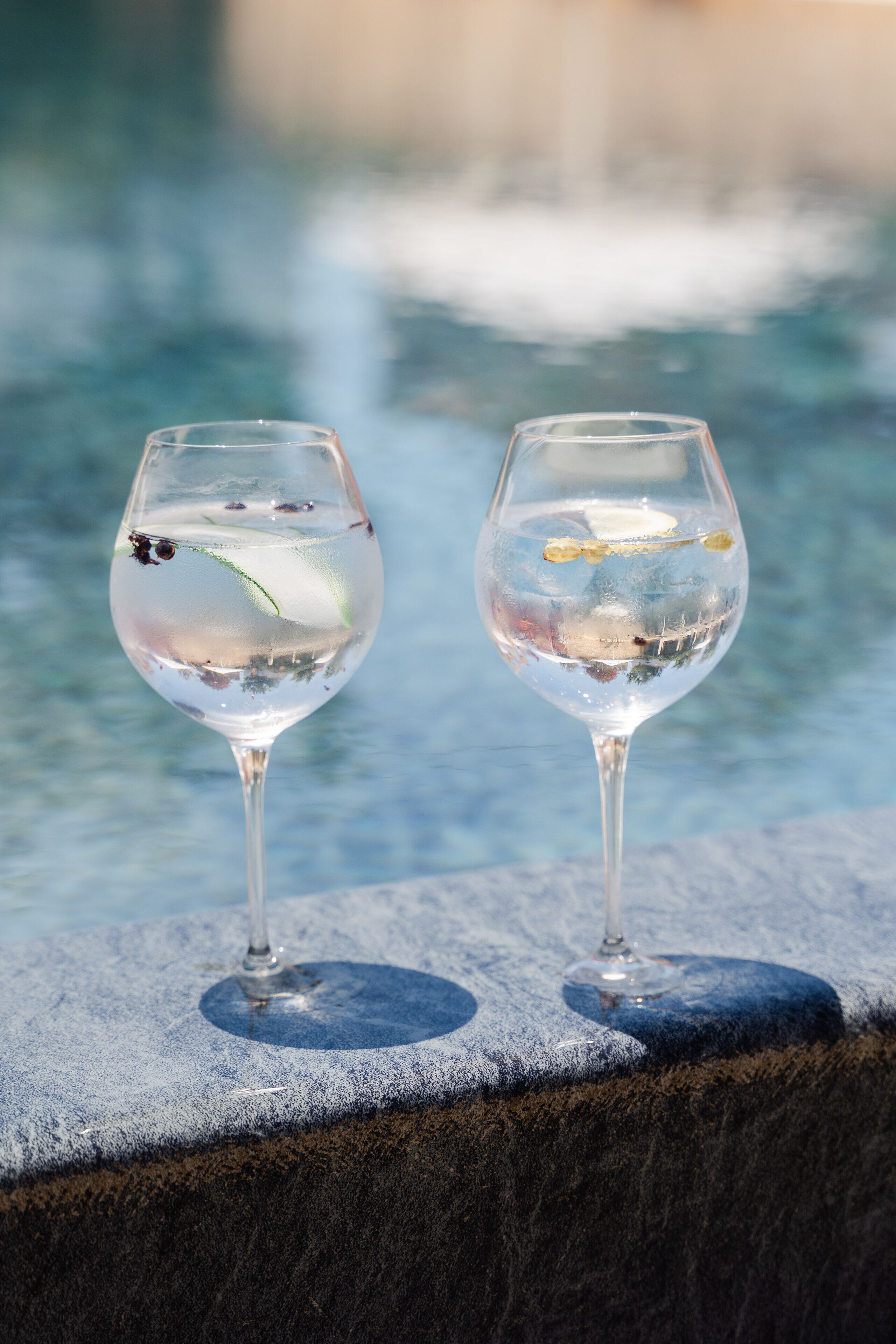 Two glasses of water with floating items on a stone ledge by a pool.