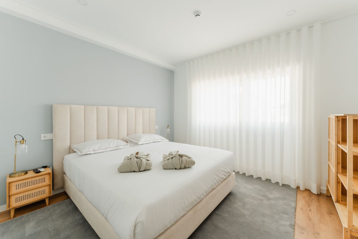 A modern bedroom with a large bed, a bedside table with a lamp, and a wooden bookshelf.