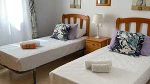 Due letti singoli con testiere in legno, biancheria da letto bianca e cuscini decorativi. Tra i letti, un comodino in legno con una lampada.