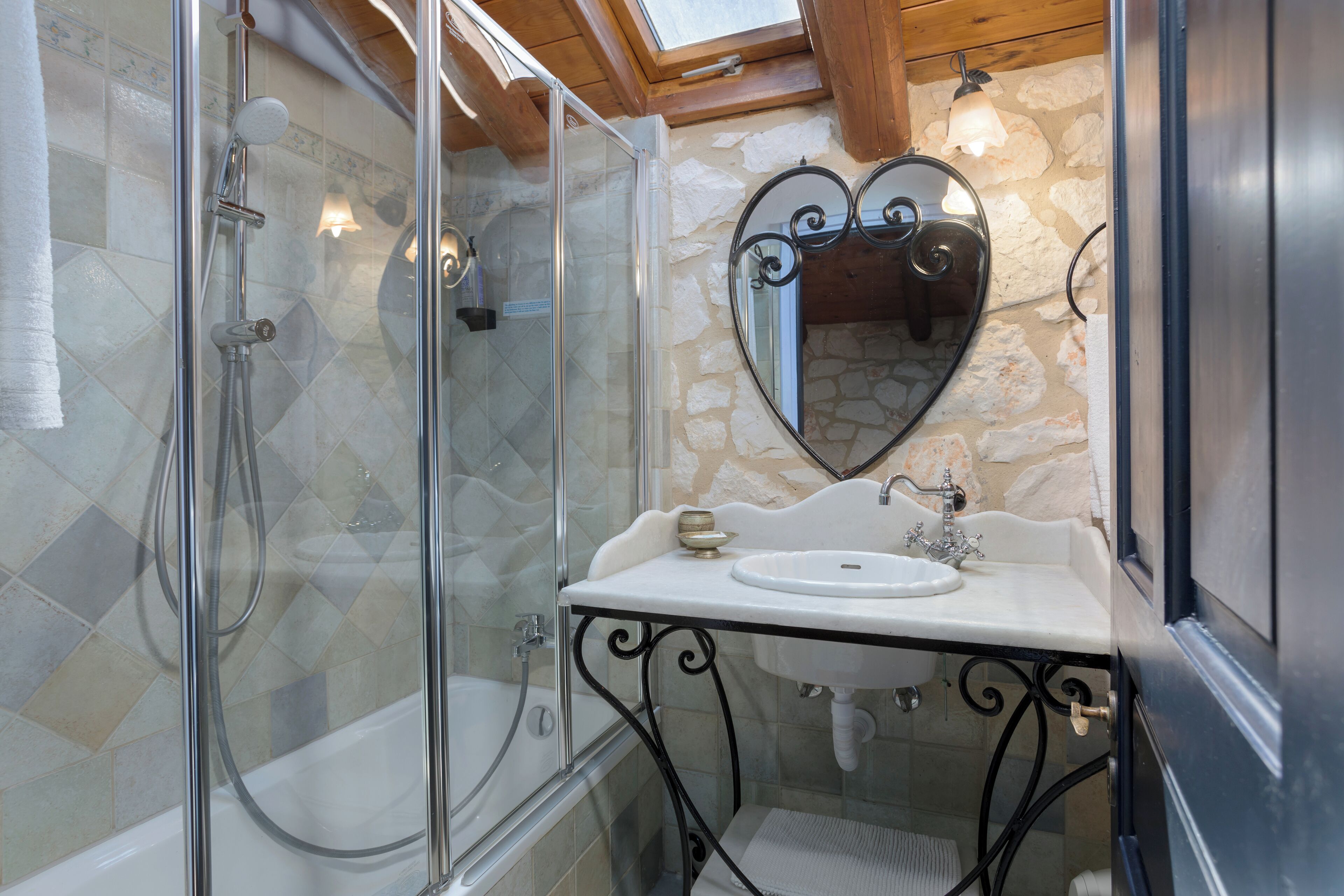 Un baño con ducha envuelta en cristal, bañera, lavabo blanco con un espejo decorativo y azulejos de piedra en la pared.