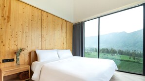A bedroom with a large bed, wooden panel walls, a nightstand, and a view of a green landscape through a large window. - ARTOTEL Cabin Bromo (Ngadisari)