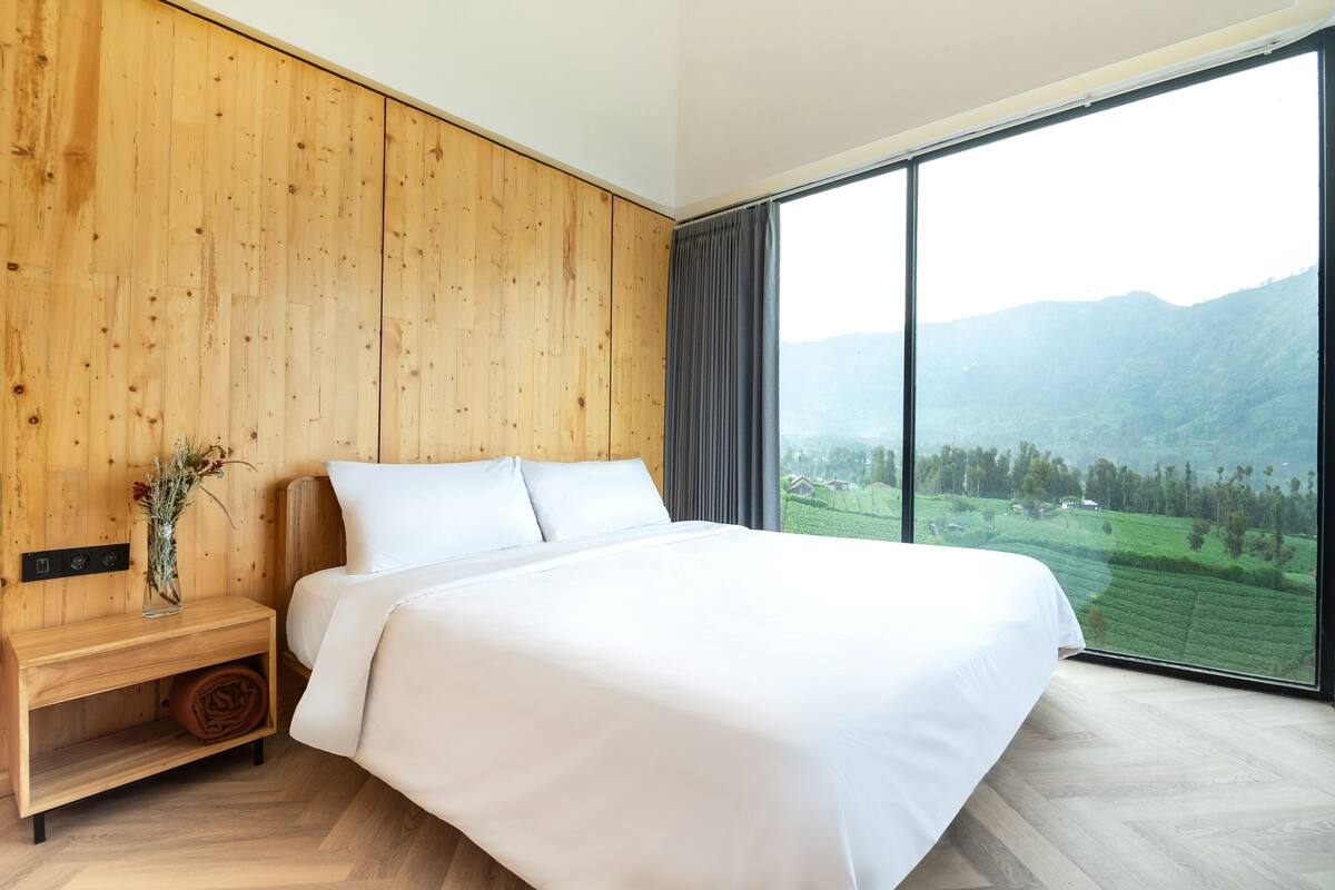 A bedroom with a large bed, wooden panel walls, a nightstand, and a view of a green landscape through a large window.