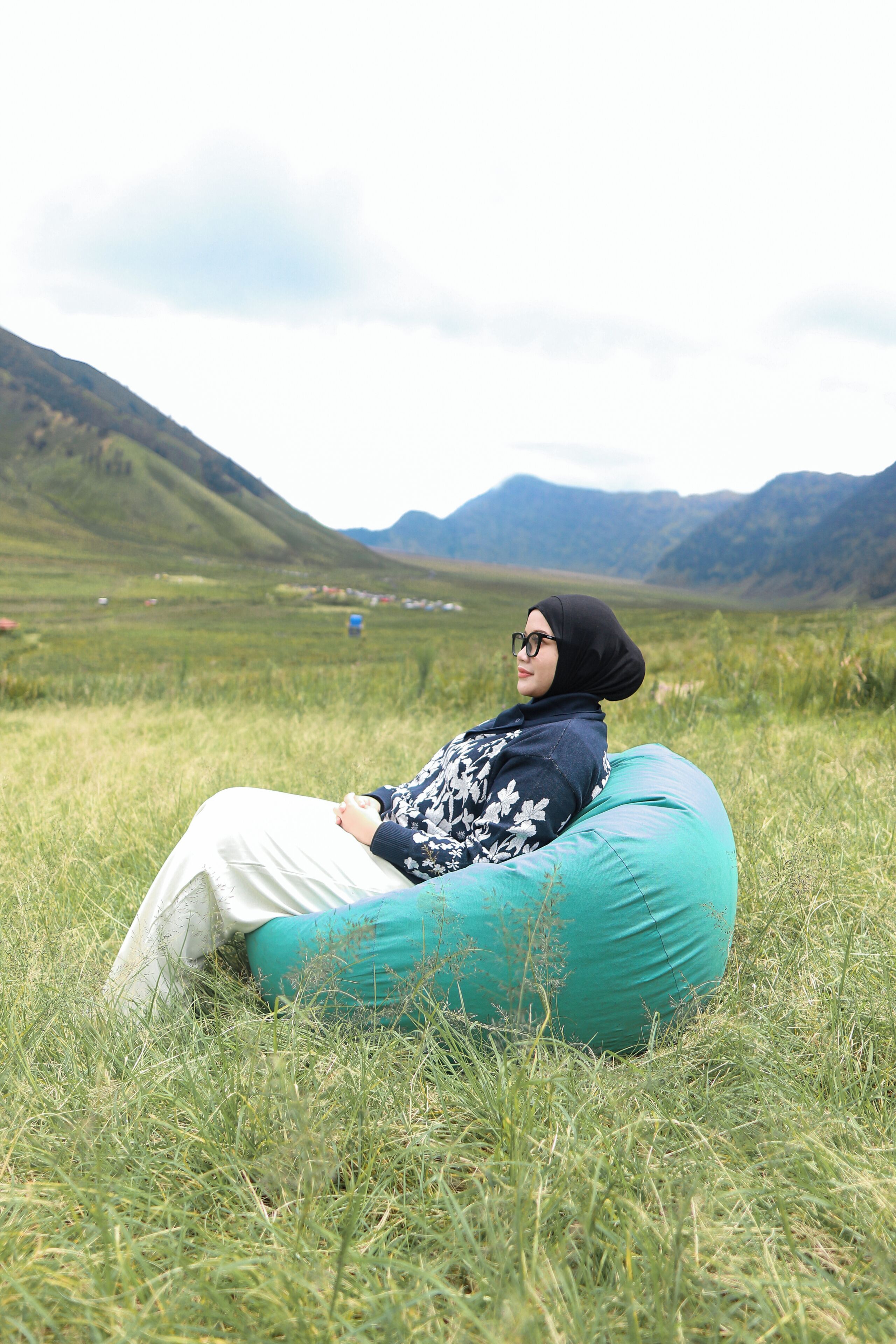 Una persona sentada en un tubo inflable verde en un campo cubierto de hierba, con montañas al fondo.