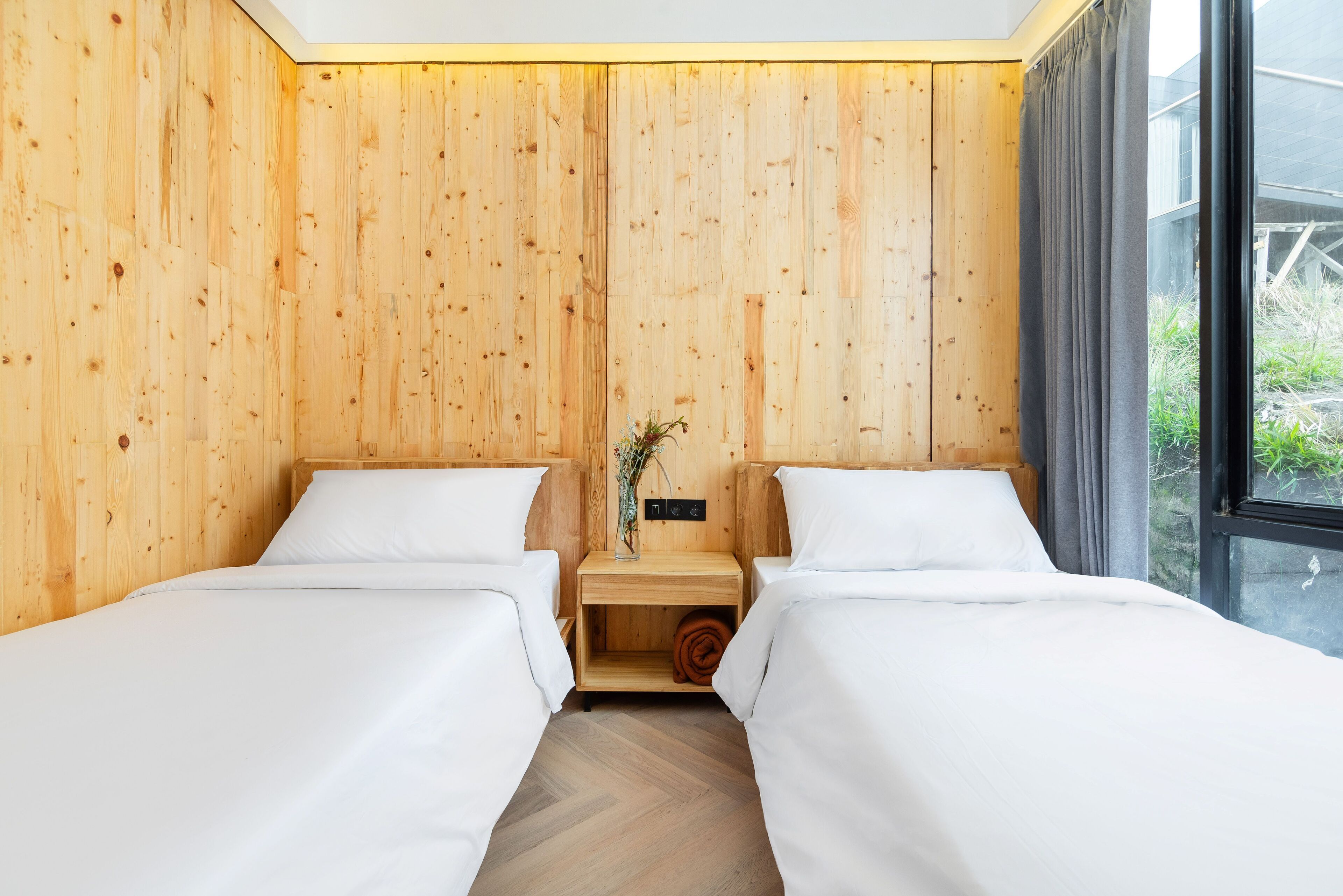 Una habitación con dos camas, paredes de madera, una mesita de noche de madera con un florero y una ventana con cortinas.