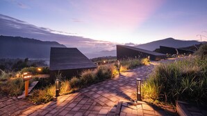 A paved pathway leading to a building with a sloped roof, surrounded by grass and plants, with a mountainous backdrop. - ARTOTEL Cabin Bromo (Ngadisari)
