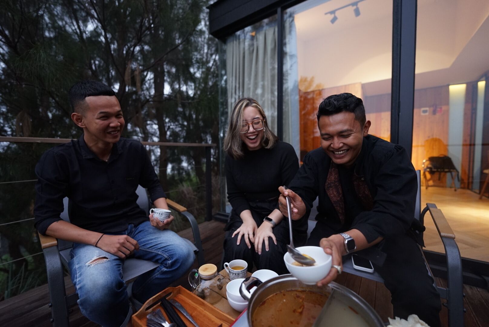 Drie personen zitten op een balkon met een tafel ervoor, koffiebekers en kommen vasthoudend.