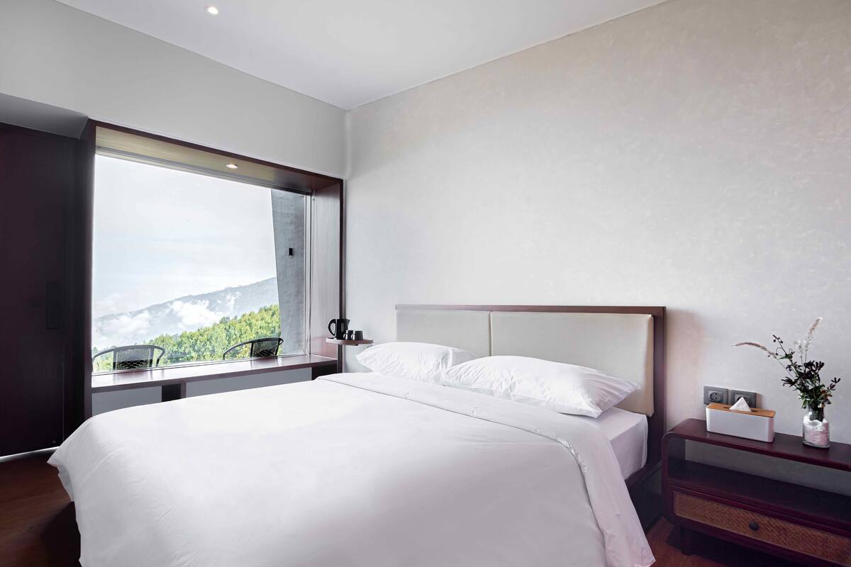 A modern hotel room with a large bed, a nightstand, a bedside table, and a window offering a mountain view.