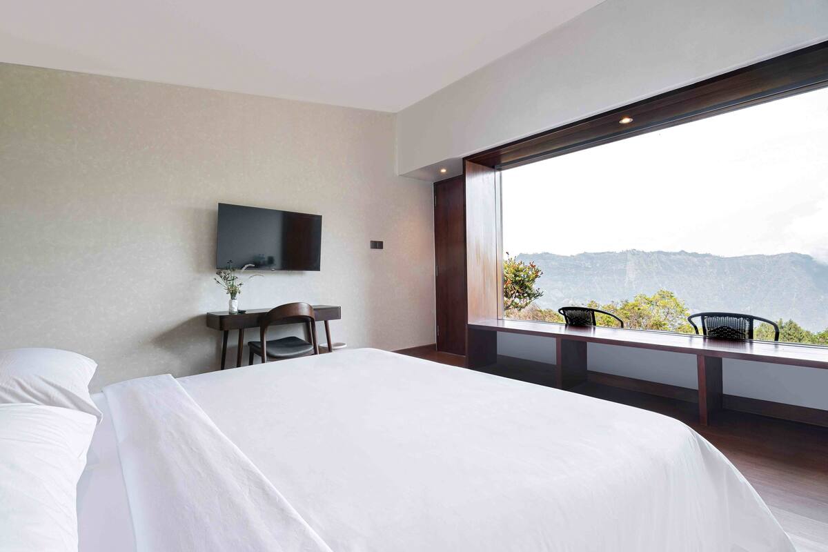 A modern hotel room with a large bed, a desk, a chair, and a TV mounted on the wall. There is a large window offering a view of mountains.