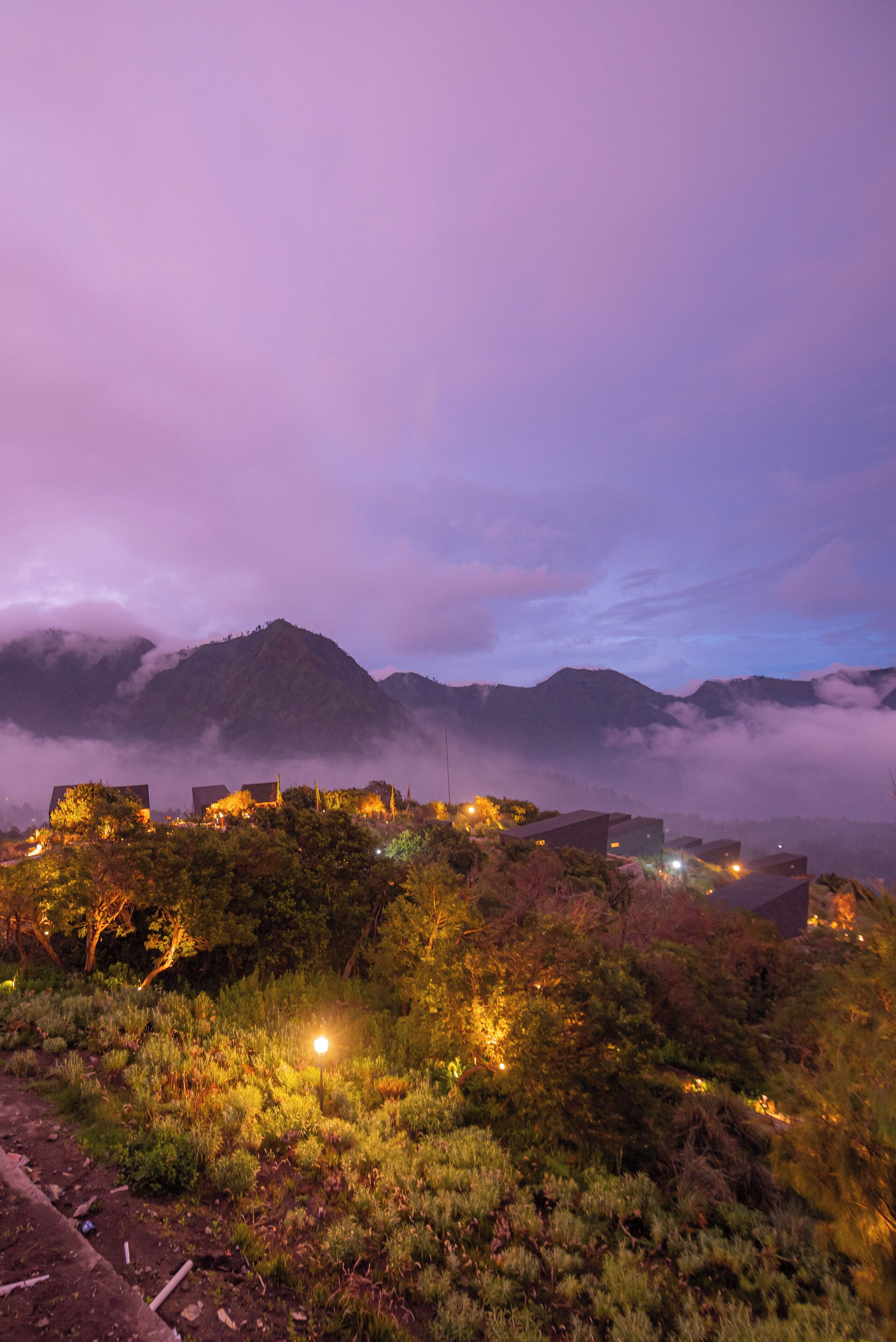 Un paisaje montañoso con edificios iluminados en primer plano, rodeado de vegetación y bajo un cielo morado.