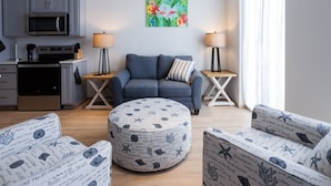 A modern living room with a grey sofa, round ottoman, and matching chairs, a kitchen with grey cabinets and a microwave, and a colorful tropical painting on the wall. - Dutch Caribbean Residence B (Oranjestad)