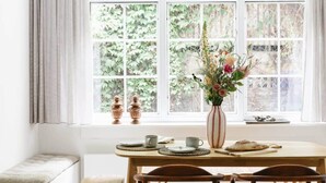 A dining area with a table set for two, a vase with flowers, and a window with a view of greenery. - 3-BR Apartment in Prime Antwerp Location (Antwerpen)