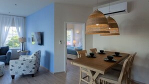 A modern living room with a wooden dining table, blue walls, and a large window with white curtains. - Dutch Caribbean Residence E (Aruba Beach Club)