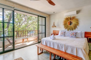 Une chambre avec une grande porte-fenêtre coulissante donnant sur un balcon, un lit avec une literie blanche, un banc en bois et un ventilateur de plafond.