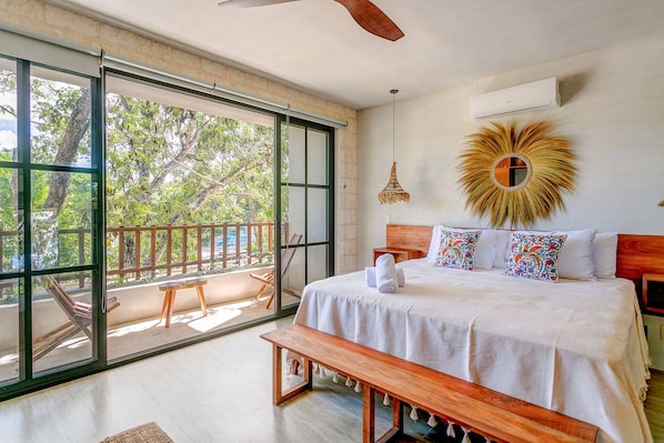 A bedroom with a large sliding glass door to a balcony, a bed with white bedding, a wooden bench, and a ceiling fan.