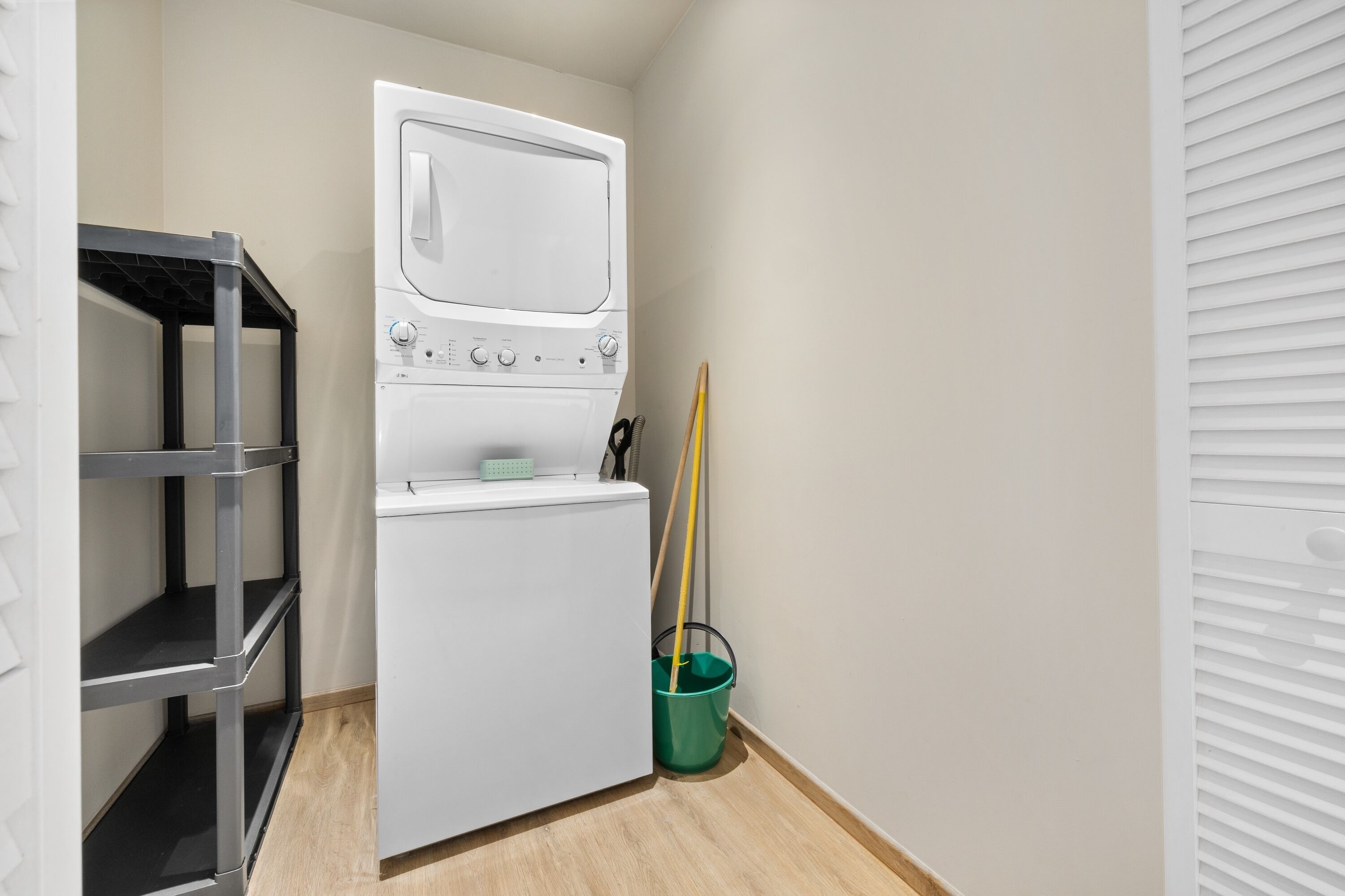 A laundry room with a stacked washer and dryer, a metal shelving unit, and a green bucket.