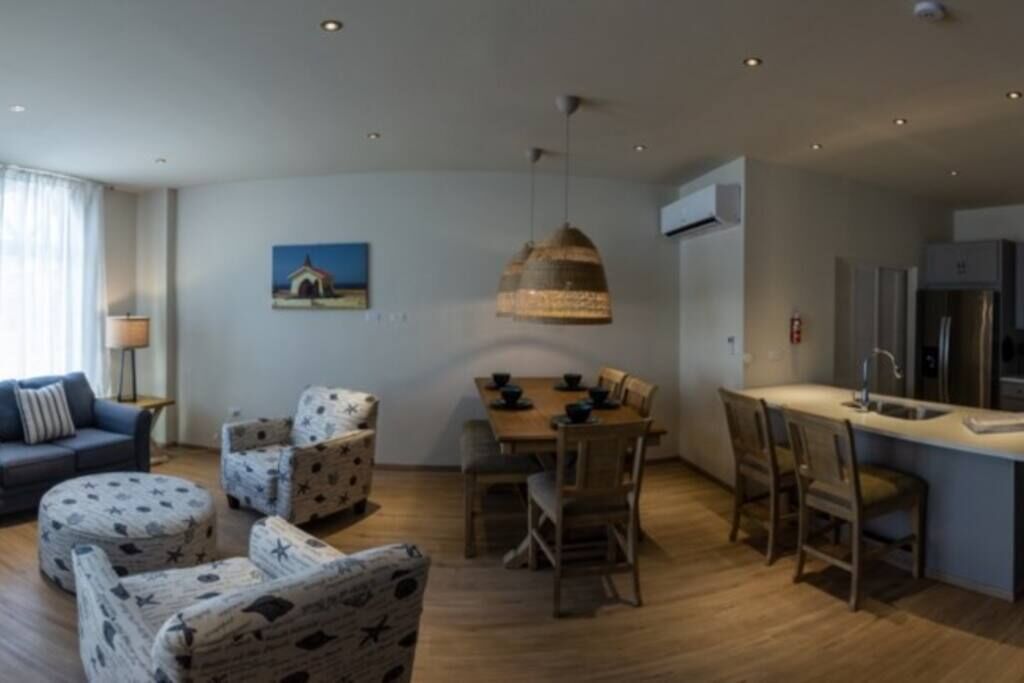 A modern living room with a sofa, armchairs, a dining table, and a kitchen area with a sink and cabinets.