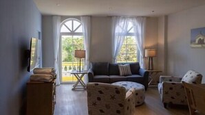 A spacious living room with a sofa, two armchairs, a small table, and a TV mounted on the wall. Large windows provide natural light and a view of the outdoors. - Dutch Caribbean Residence D (Oranjestad)
