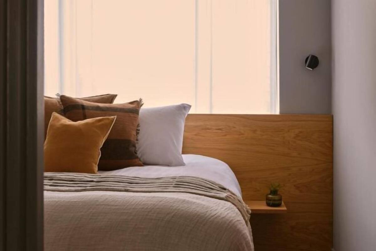 A neatly made bed with a striped blanket and two pillows, a small wooden nightstand with a plant, and a window with blinds.