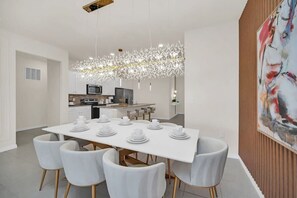 Un moderno comedor con una mesa blanca dispuesta para seis personas, una cocina al fondo y un cuadro abstracto grande en la pared.