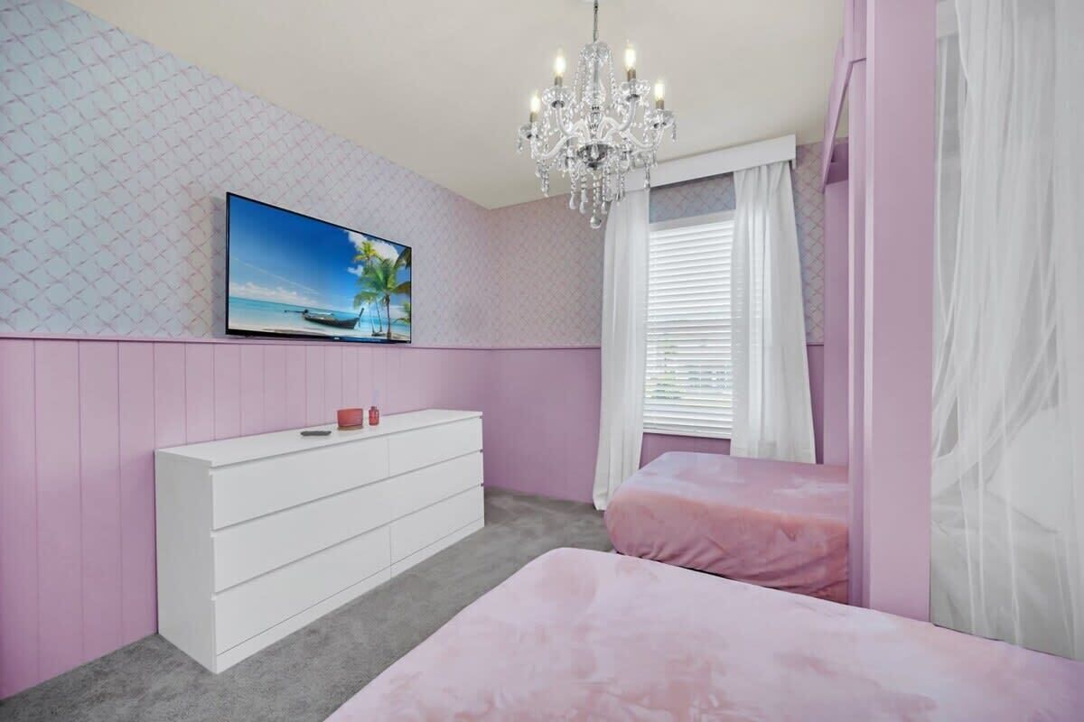 A bedroom with a pink theme, featuring a white dresser, a bed with pink bedding, a TV on the wall, and a chandelier.