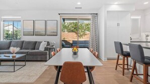 A modern living room with a sofa, coffee table, and a dining area with chairs and a table. There is a view of the outdoors through a sliding door. - Desert Hideaway: 2BR/2BA, BBQ, FirePit, PGA West! (La Quinta)