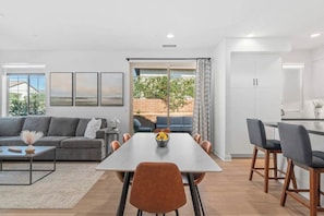A modern living room with a sofa, coffee table, and a dining area with chairs and a table. There is a view of the outdoors through a sliding door. - Desert Hideaway: 2BR/2BA, BBQ, FirePit, PGA West! (La Quinta)